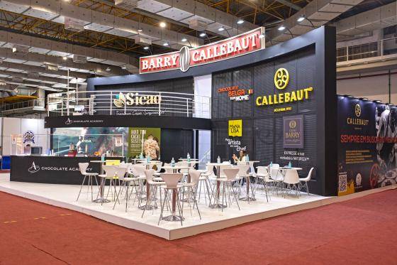 foto panoramica do stande da Barry Callebaut na FIPAN 2022 com dois andares, em sua maioria na cor preta com o logo de todas as marca. Na frente cadeiras e mesas brancas